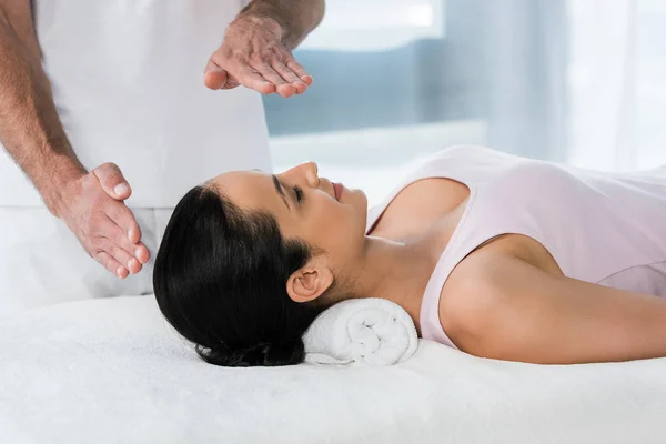 Vue recadrée du guérisseur mettant les mains au-dessus de la tête de la fille brune sur la table de massage — Photo de stock