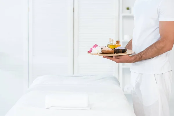 Cropped view of man holding tray with bottles of oil, bowls with sea salt and flowers in spa center — Stock Photo