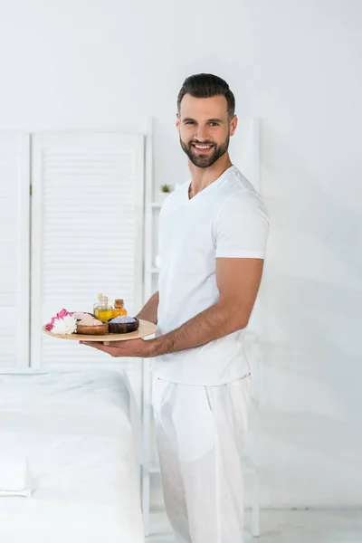 Happy bearded man holding tray with bottles of oil, bowls with sea salt and flowers in spa center — Stock Photo