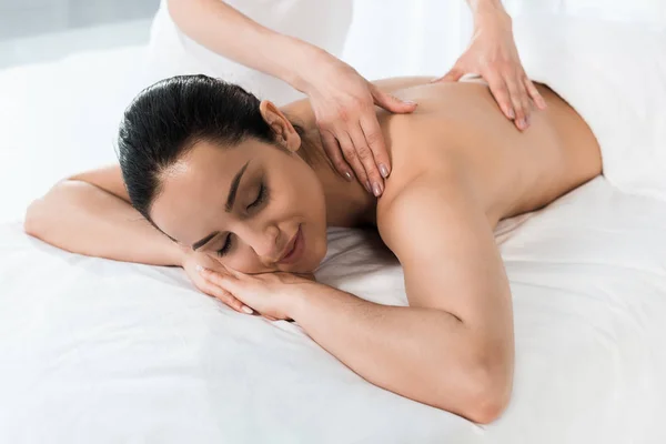 Recortado vista de masajista haciendo masaje a feliz morena mujer acostada en mesa de masaje - foto de stock