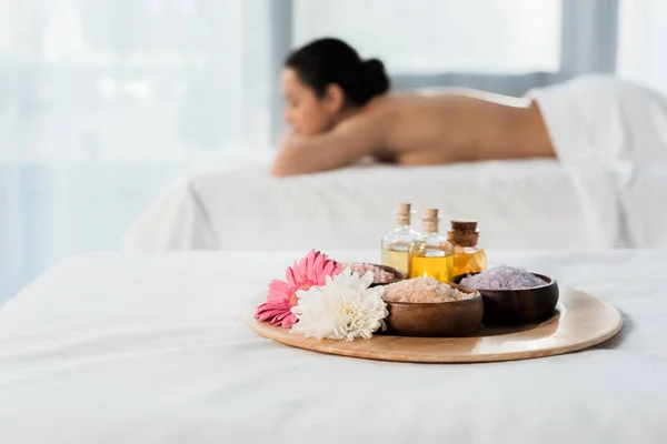 Selective focus of tray with bottles of oil, bowls with sea salt and flowers near brunette woman in spa center — Stock Photo