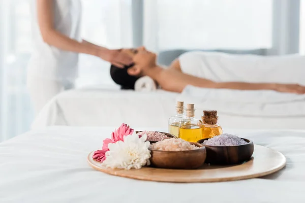 Selektiver Fokus des Tabletts mit Ölflaschen, Schalen mit Meersalz und Blumen in der Nähe von Frauen im Kurzentrum — Stockfoto