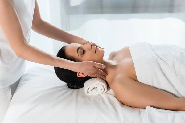 Recortada vista de masajista haciendo masaje de cuello a atractiva morena tumbada en mesa de masaje - foto de stock