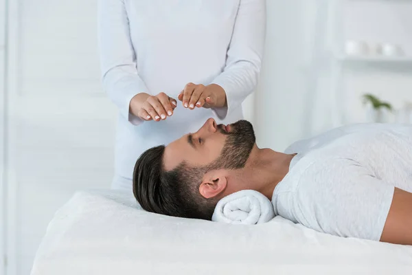 Visão cortada de curandeiro colocando as mãos acima da cabeça de homem bonito na mesa de massagem — Fotografia de Stock