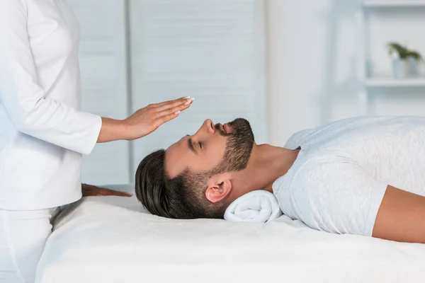 Vue recadrée du guérisseur mettant les mains au-dessus de la tête du bel homme sur la table de massage — Photo de stock