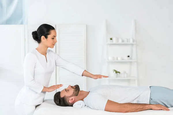 Curandeiro atraente colocando as mãos acima do corpo de homem bonito na mesa de massagem — Fotografia de Stock