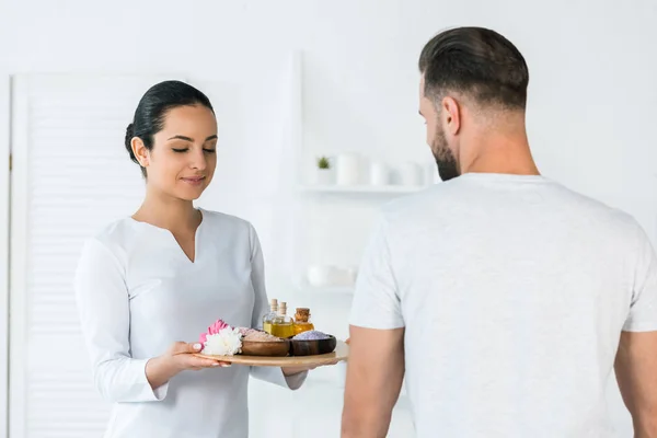 Mulher atraente segurando bandeja com garrafas de óleo, tigelas com sal marinho e flores perto do homem no centro de spa — Stock Photo