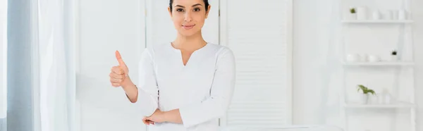 Plano panorámico de atractivo masajista de pie en el centro de spa y mostrando el pulgar hacia arriba - foto de stock