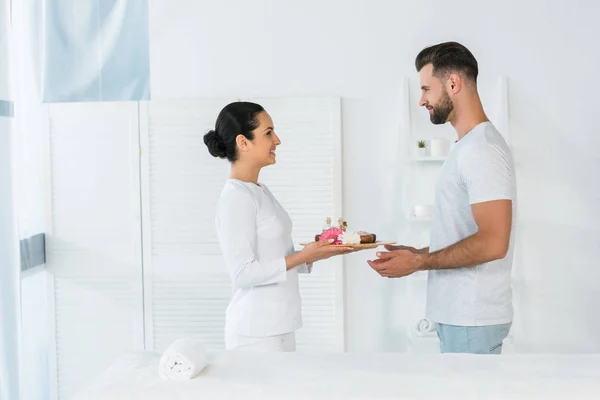 Beautiful brunette masseur holding tray with bottles of oil, bowls with sea salt and flowers near man in spa center — Stock Photo