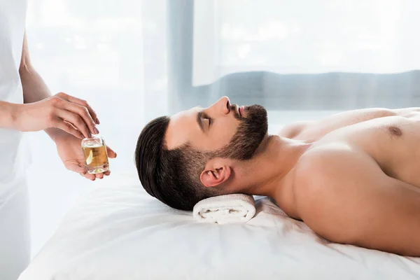 Vista recortada de masajista sosteniendo botella con aceite cerca de hombre guapo con los ojos cerrados - foto de stock