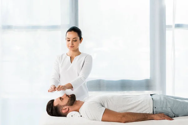 Attractive healer putting hands above head while healing handsome bearded man — Stock Photo