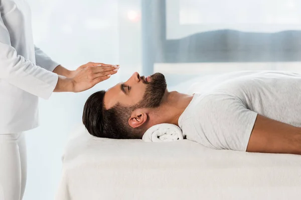 Vue recadrée du guérisseur mettant les mains au-dessus de la tête de l'homme barbu avec les yeux fermés — Photo de stock