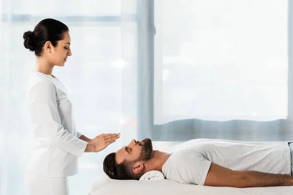 Attractive brunette healer putting hands above head of bearded man with closed eyes — Stock Photo