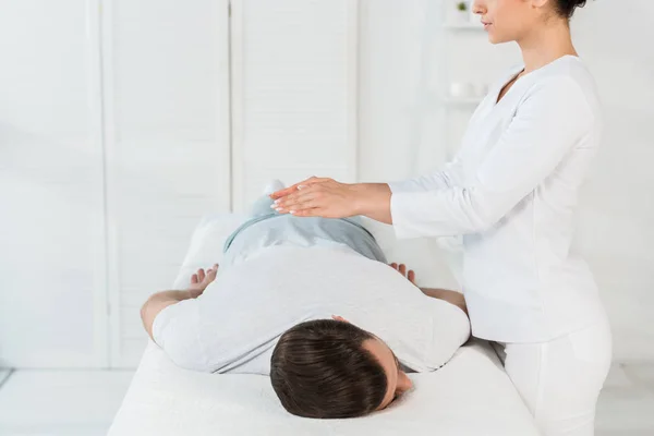 Vista ritagliata del guaritore mettendo le mani sopra la schiena dell'uomo sul tavolo da massaggio — Foto stock