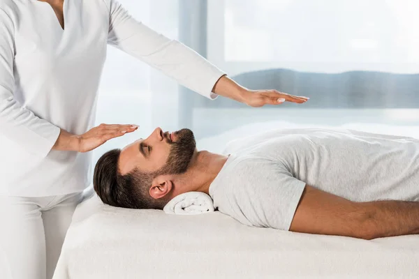 Vista recortada de curandero poniendo las manos por encima del cuerpo del hombre barbudo - foto de stock