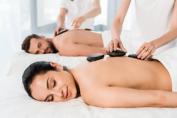 Selective focus of masseur doing hot stone massage to attractive woman with closed eyes near bearded man — Stock Photo