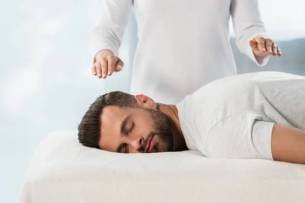 Vista recortada del sanador con las manos por encima del cuerpo del hombre en la mesa de masaje - foto de stock