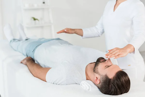 Vista recortada del sanador con las manos por encima del cuerpo del hombre descansando sobre la mesa de masaje - foto de stock