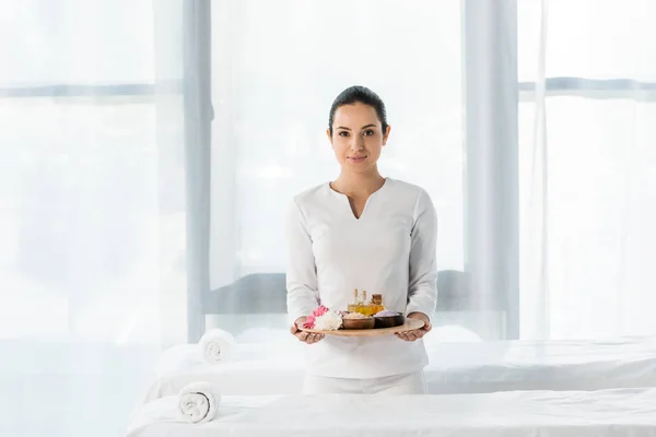 Attractive brunette masseur holding tray with bottles of oil, bowls with sea salt and flowers in spa center — Stock Photo