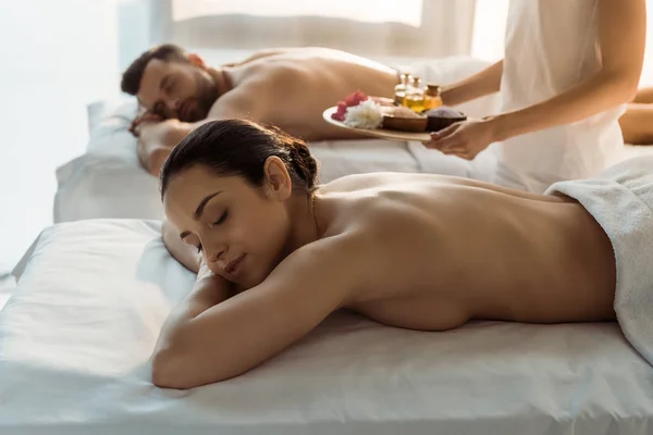 Selective focus of masseur holding tray with sea salt and bottles of oil near woman and man in spa center — Stock Photo