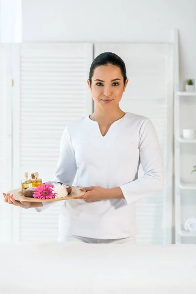 Attrayant masseur tenant plateau avec des bouteilles d'huile, sel de mer et fleurs — Photo de stock