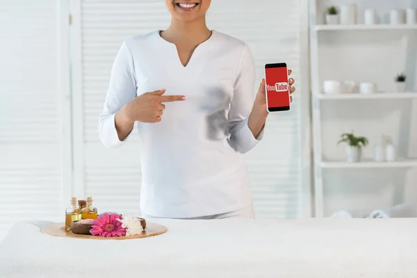 KYIV, UKRAINE - MAY 20, 2019: cropped view of cheerful masseur pointing with finger at smartphone with youtube app near tray with bottles of oil and flowers — Stock Photo