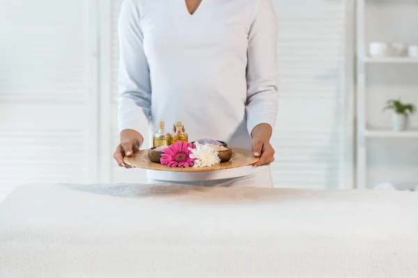 Vue recadrée du jeune masseur tenant un plateau avec des bouteilles d'huile, de sel de mer et de fleurs — Photo de stock