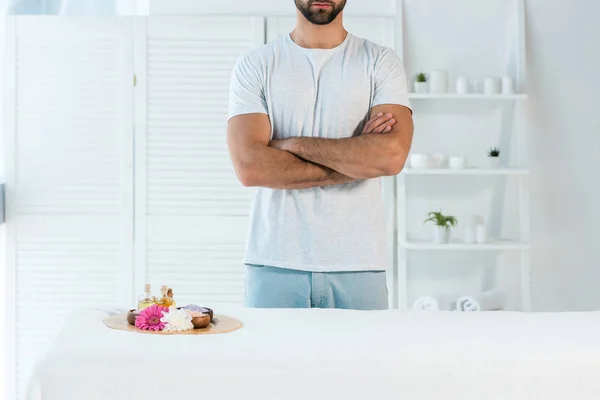 Vista cortada do homem de pé com braços cruzados perto da bandeja com garrafas de óleo, sal marinho e flores — Fotografia de Stock