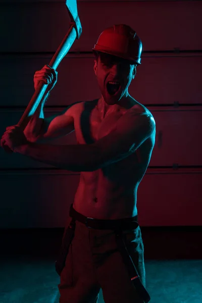 Screaming sexy shirtless fireman in helmet holding flat head axe in darkness — Stock Photo