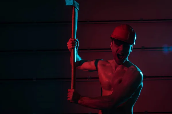 Screaming sexy shirtless fireman in helmet holding flat head axe in darkness — Stock Photo