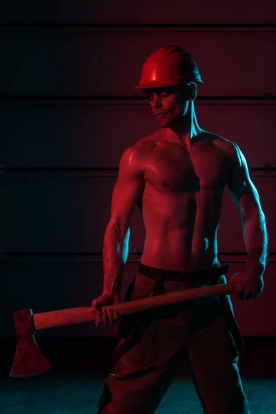 Sexy shirtless fireman in protective helmet holding flat head axe in darkness — Stock Photo