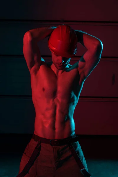 Sexy shirtless fireman in protective helmet posing with hands up in darkness — Stock Photo