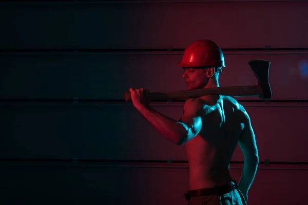 Sexy shirtless fireman in protective helmet holding flat head axe in darkness — Stock Photo