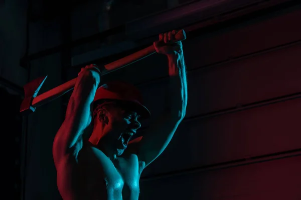 Sexy sin camisa bombero en casco con la cabeza plana hacha y gritando en la oscuridad - foto de stock