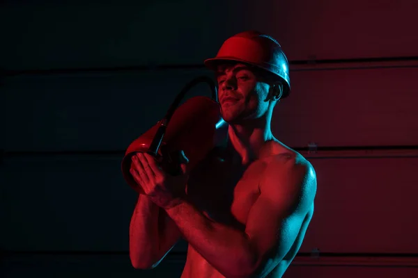 Sexy shirtless fireman in protective helmet holding fire extinguisher in darkness — Stock Photo