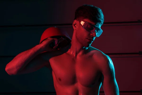 Sexy bombero sin camisa en gafas protectoras sosteniendo hardhat en la oscuridad - foto de stock