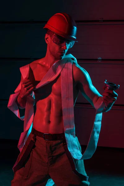 Sexy shirtless fireman in protective helmet holding fire hose in darkness — Stock Photo