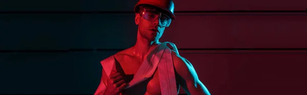 Panoramic shot of sexy fireman in protective equipment holding fire hose in darkness — Stock Photo