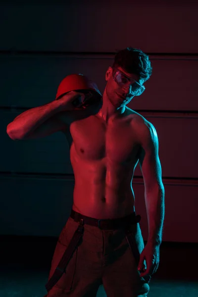 Sexy shirtless fireman in protective goggles holding hardhat in darkness — Stock Photo