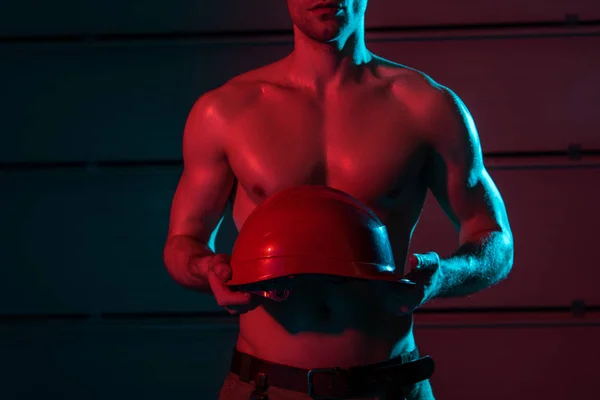 Cropped view of sexy shirtless fireman holding protective helmet in darkness — Stock Photo