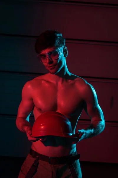 Sexy shirtless fireman in protective goggles holding hardhat in darkness — Stock Photo