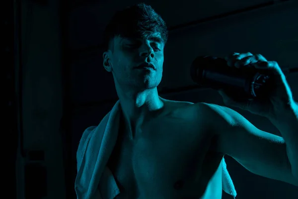 Sexy uomo senza maglietta con asciugamano in possesso di bottiglia di sport nel buio — Foto stock