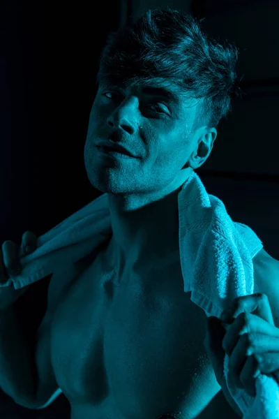 Улыбающийся сексуальный мужчина с полотенцем, смотрящий на камеру в темноте — стоковое фото