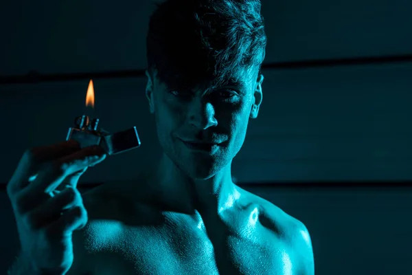 Sexy shirtless muscular man holding lighter and smiling in darkness — Stock Photo