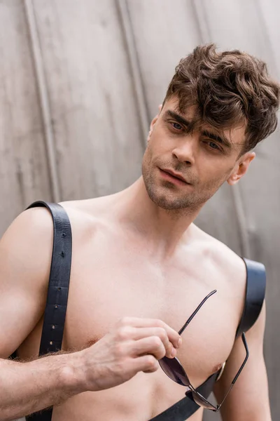 Curious shirtless sexy man in sword belt holding sunglasses and looking at camera — Stock Photo