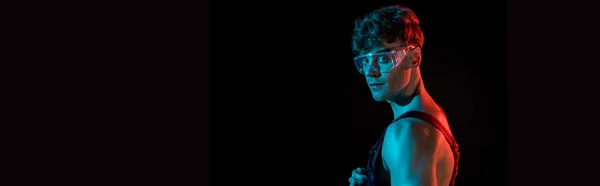 Panoramic shot of sexy fireman in overall and protective goggles isolated on black — Stock Photo