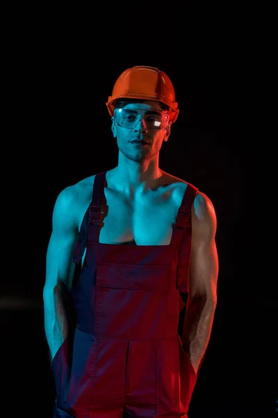 Sexy fireman in overall, hardhat and protective goggles with hands in pockets isolated on black — Stock Photo
