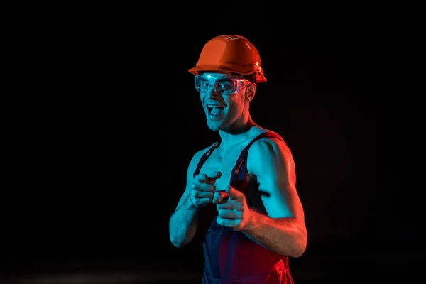 Lächelnder sexy Feuerwehrmann in Overall, Schutzbrille und Schutzbrille, die mit Fingern auf Schwarz zeigt — Stockfoto