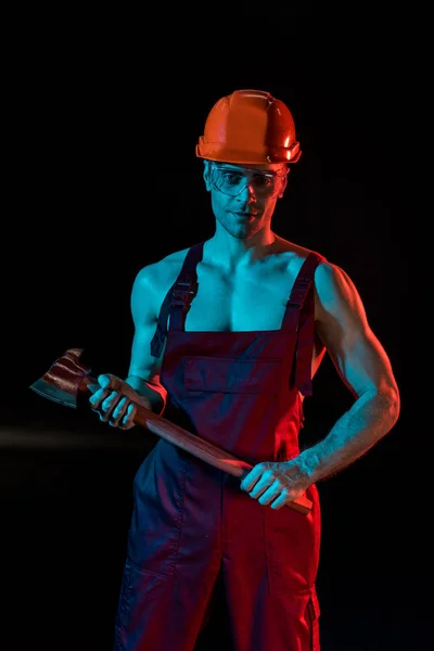 Sexy fireman in overall, hardhat and protective goggles holding flat head axe on black — Stock Photo