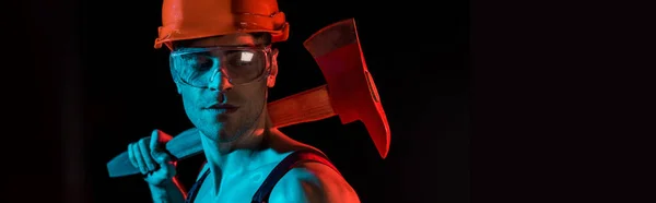 Panoramic shot of shirtless fireman in hardhat and protective goggles holding flat head axe isolated on black — Stock Photo